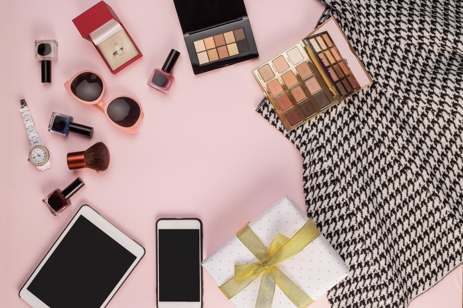 A flat lay of a scarf, a tablet, a smartphone, a watch, sunglasses, a makeup palette, a makeup brush, nail polishes, a ring box, and a gift box.